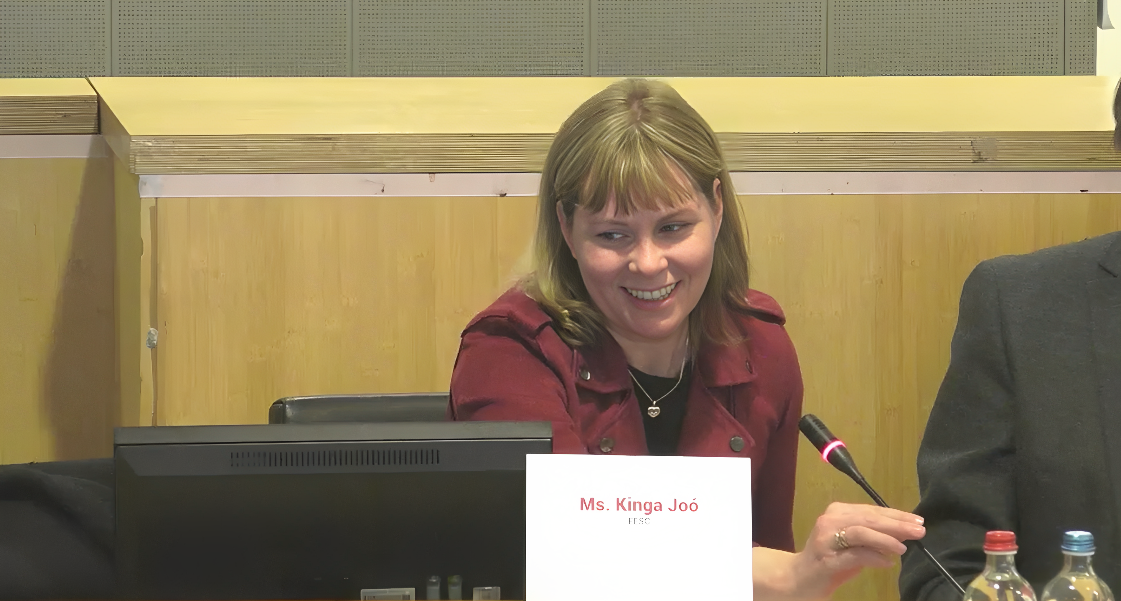 Ms. Kinga Joó wearing a smile as she sits at the speaker's table, confidently holding the microphone.